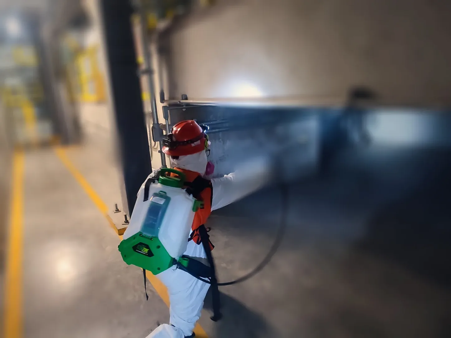 Técnico con casco y aspersor de mochila aplicando junto a un muro en una planta